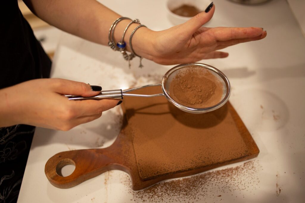 Les mains d'une femme tamisent de la poudre de cacao sur une planche à découper en bois dans une cuisine.