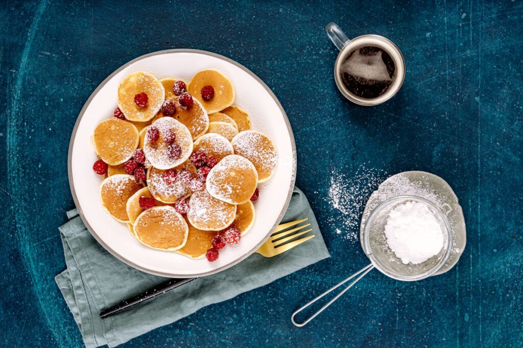 Vue aérienne de mini crêpes avec des framboises, du sucre glace et du café sur un fond bleu.