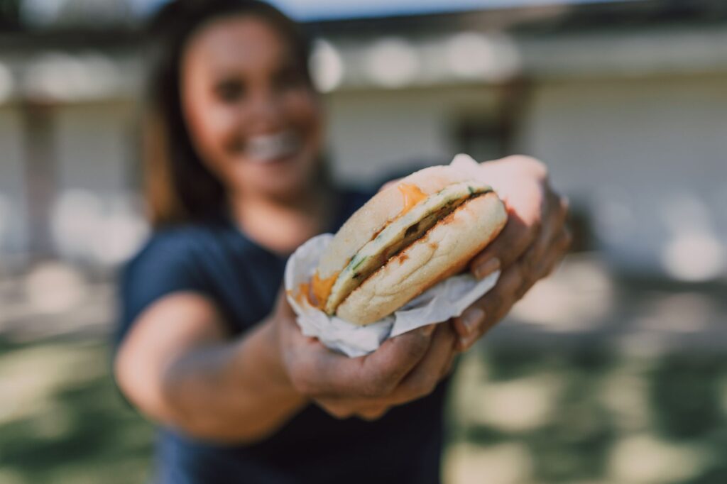 Une femme joyeuse offrant un sandwich savoureux dans un cadre extérieur ensoleillé.