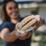 Une femme joyeuse offrant un sandwich savoureux dans un cadre extérieur ensoleillé.