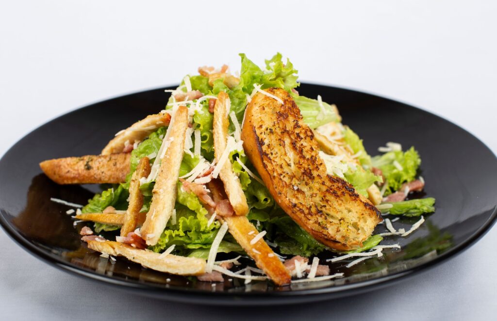 Gros plan sur une salade César avec des croûtons, du Parmesan et des légumes frais dans une assiette noire.