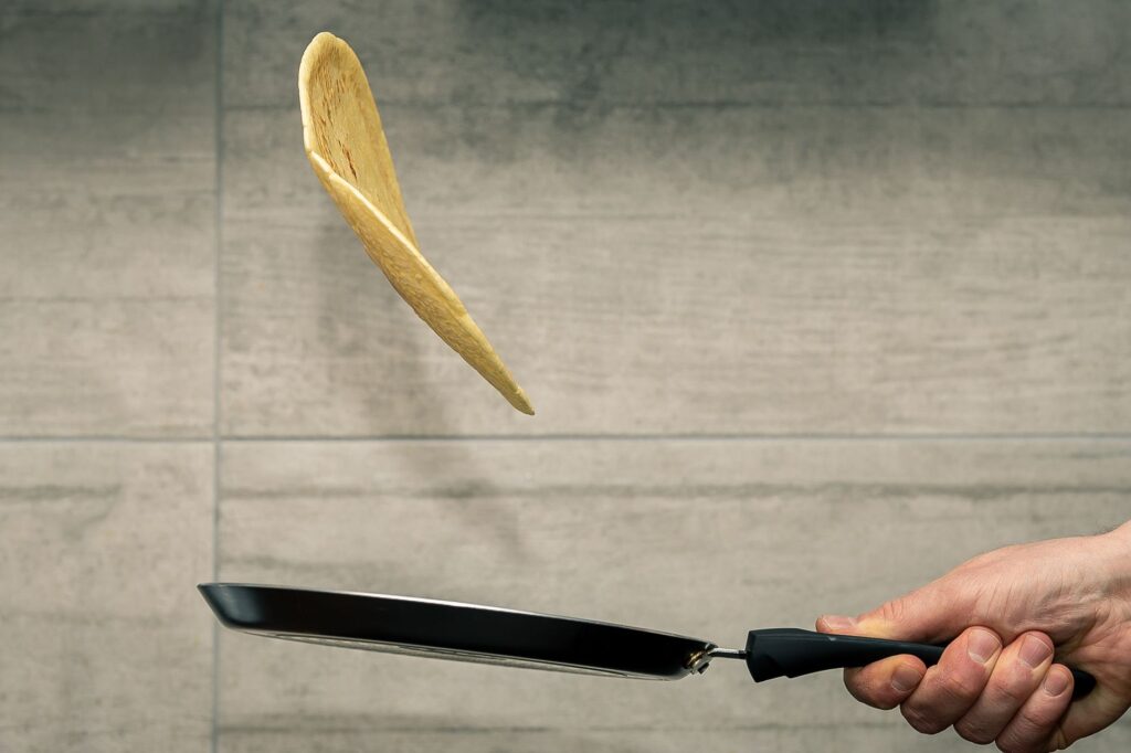 Une main retourne habilement une crêpe en plein vol dans une poêle, mettant en valeur l'expertise culinaire.