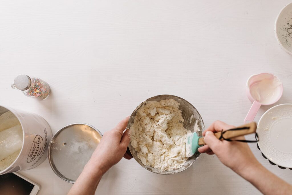Une personne mélangeant de la pâte maison dans un bol avec des ustensiles de pâtisserie sur une table blanche.