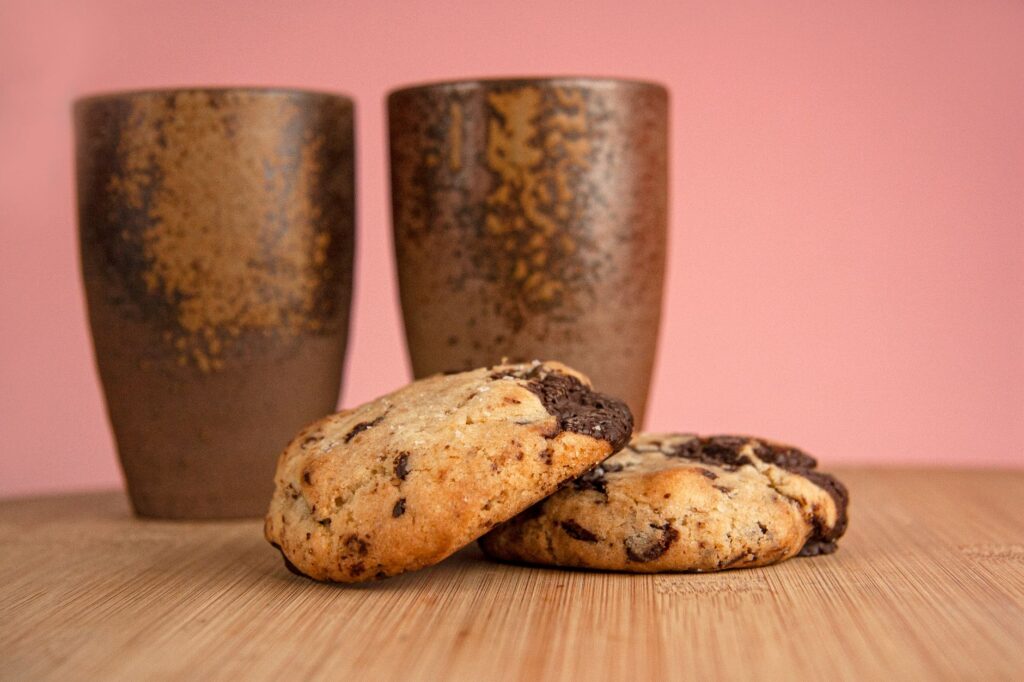 Gros plan sur des biscuits aux pépites de chocolat faits maison avec des tasses rustiques sur un fond rose.