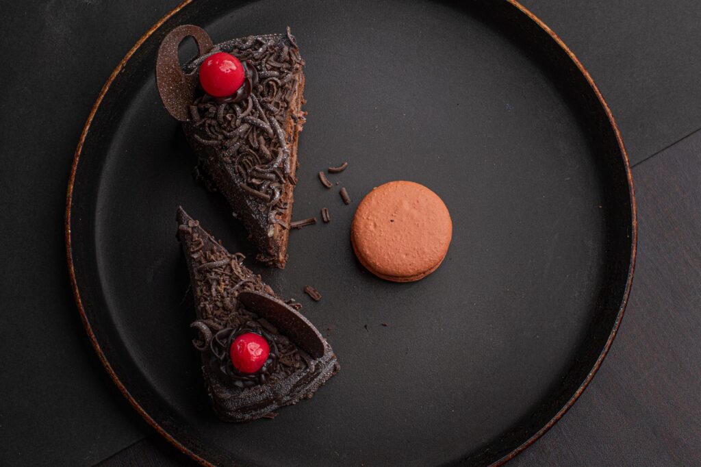 Un gros plan de parts de gâteau au chocolat avec des cerises et un macaron sur une assiette noire.