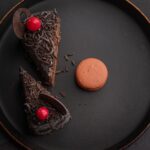 Un gros plan de parts de gâteau au chocolat avec des cerises et un macaron sur une assiette noire.