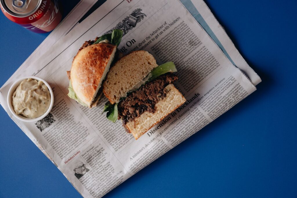 Vue plongeante d'un délicieux burger accompagné de cola et de sauce servi sur un journal sur une table bleue.