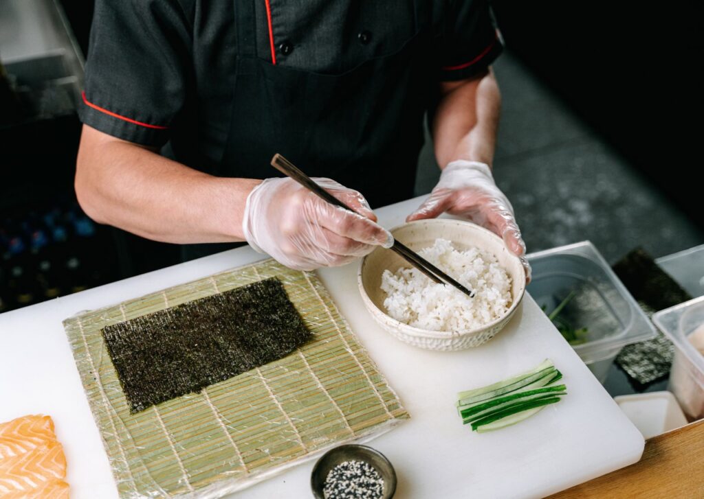 Gros plan d'un chef préparant des sushis avec des baguettes, du riz et du nori sur une planche à découper.